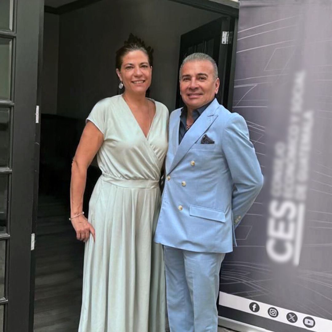 A darf-haired woman in a light-grey long dress, a man in a light-blue suit with grey hair on her right. 